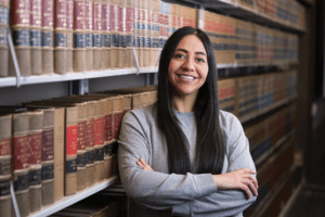 Heidi Chamorro-Leon, a Hispanic woman with dark hair and dark eyes, stands in front of bookshelf