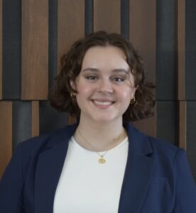 Cait McKee, a young white woman with short, curly brown hair wearing a white blouse and navy blazer